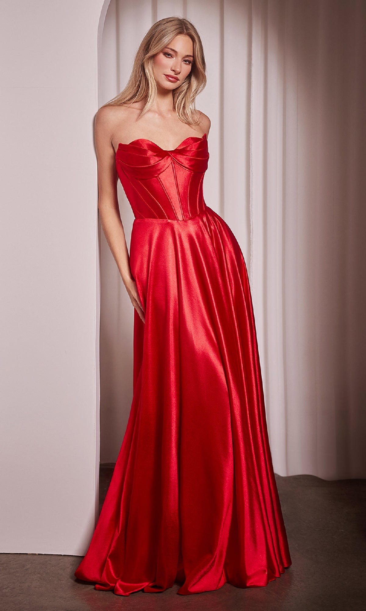 Woman modeling the Ladivine CD378 Strapless Long A-Line Prom Dress with a corset-style bodice and flowing red satin skirt, standing indoors against a white curtain backdrop.